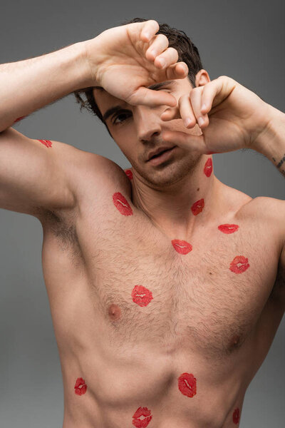 sexy shirtless man with red lipstick prints on torso obscuring face with hands while looking at camera isolated on grey