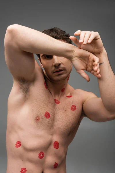 sexy man with red lipstick prints on muscular torso obscuring face and looking at camera isolated on grey