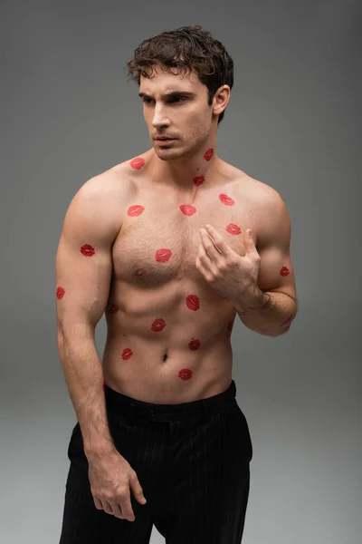 brunette muscular man with red lipstick prints on shirtless body looking away on grey background