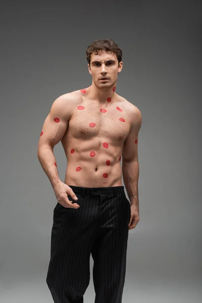 sexy man with red lipstick marks on muscular body looking at camera on grey background
