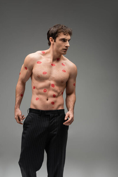 sexy man in black pants posing with red kiss prints on shirtless muscular torso and looking away on grey background