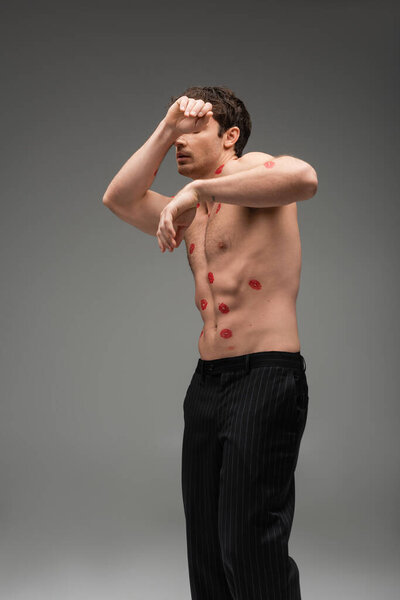 man in black pants with red lip prints on shirtless torso obscuring face while posing on grey background
