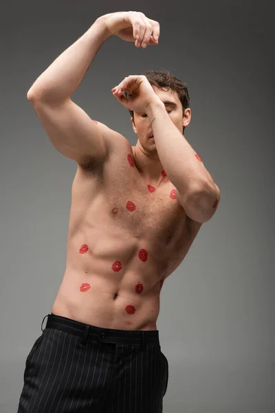 sexy man with red kiss prints on muscular body obscuring face with raised hands isolated on grey