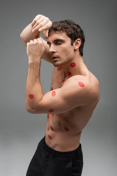 shirtless muscular man with red kiss prints on body posing with closed eyes and hands near face on grey background