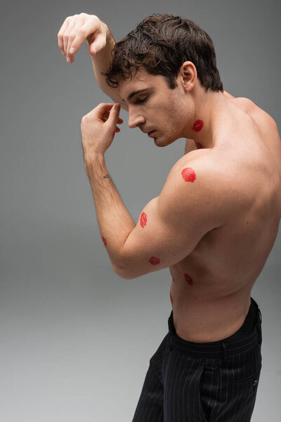 side view of brunette man with red lipstick marks on shirtless body posing on grey background