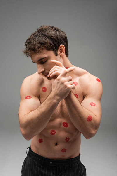 brunette man with red lipstick marks on shirtless muscular body posing with closed eyes on grey background