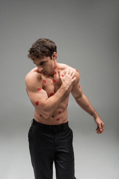 athletic man in black trousers posing with red lip prints on muscular torso on grey background
