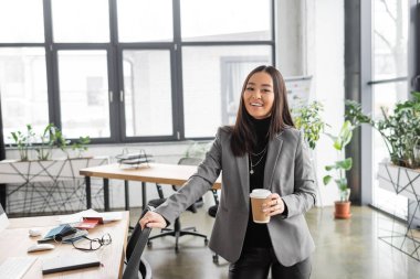 Positive asian interior designer holding coffee to go and looking at camera in office 