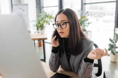 Brunette asian interior designer talking on smartphone and looking at computer in studio 