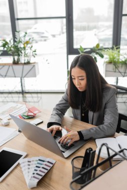 Asian designer using laptop near devices and color samples in studio 