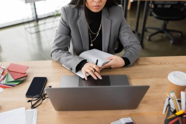 Cropped view of interior designer holding notebook near devices and color samples in office 