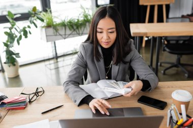Asian designer holding color samples and using laptop in office 