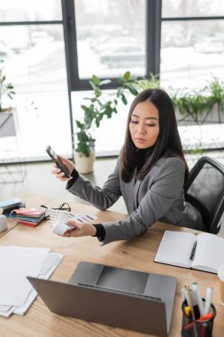 Asian designer looking at color samples and using smartphone in studio 