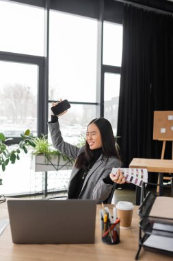 Cheerful asian interior designer holding cellphone and holding color swatches in studio 