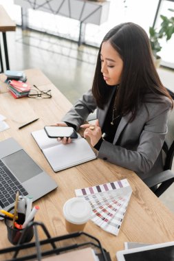 Asian designer using mobile phone near coffee to go and color samples in office 