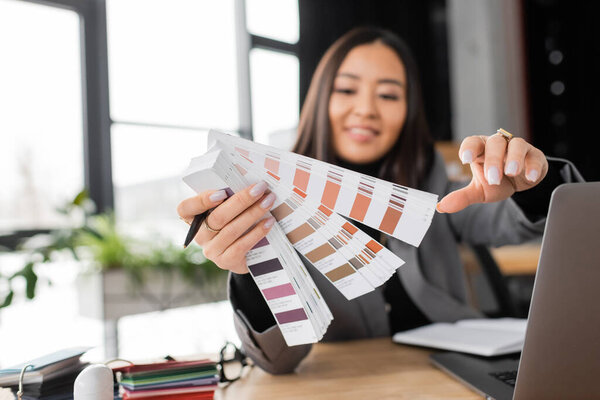 Blurred asian interior designer holding color samples near laptop in office 