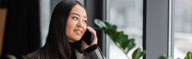 Positive asian designer talking on mobile phone in office, banner 