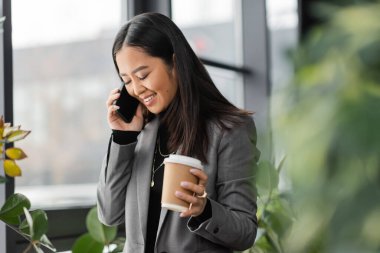 Positive asian designer talking on mobile phone and holding takeaway coffee in office  clipart