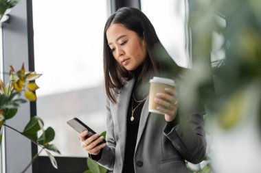 Brunette asian interior designer using smartphone and holding coffee in paper cup near plants in office 