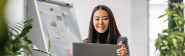 Positive asian designer holding laptop near plants in studio, banner 