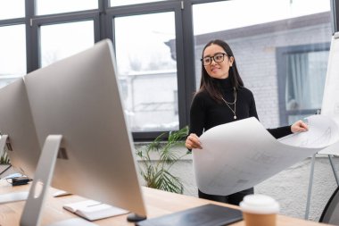 Smiling asian interior designer holding blueprint and looking at computer monitor in studio 