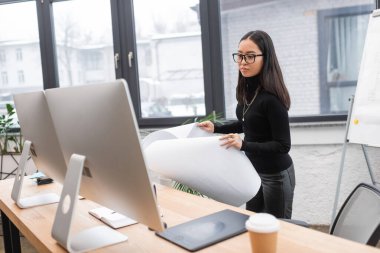 Asian interior designer holding blueprint near computers in office 