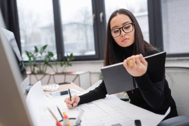 Brunette asian designer holding notebook near blueprint and computers on office 