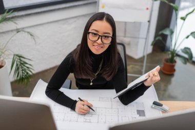 Cheerful asian interior designer holding notebook and looking at camera near blueprint in office  clipart