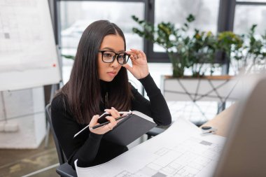 Young asian interior designer in eyeglasses holding notebook near computer and blueprint in office 