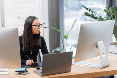 Asian interior designer using graphic tablet near computers and devices in office  clipart