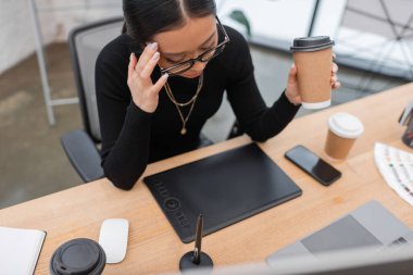 Overhead view of tired asian interior designer holding coffee near devices in studio 