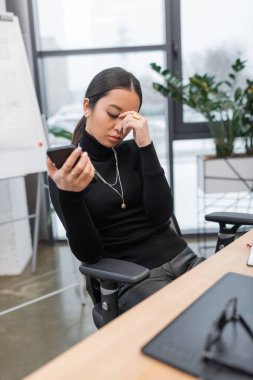 Exhausted asian interior designer holding smartphone in studio 