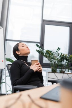 Asian designer holding coffee to go and relaxing near working table in office  clipart