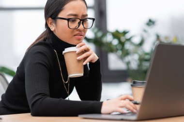 Upset asian designer holding paper cup and using laptop in office 