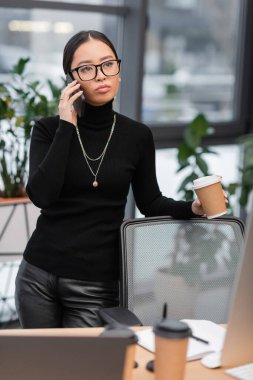 Asian interior designer talking on smartphone and holding coffee to go near devices in office 