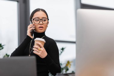 Asian designer talking on smartphone and holding coffee near computers in office  clipart
