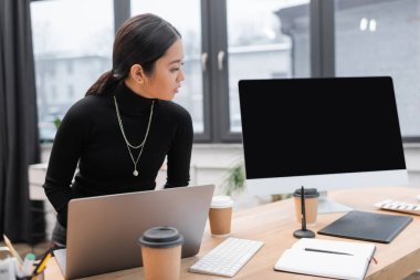 Asian interior designer using laptop near graphic tablet and computer monitor in office  clipart