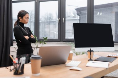 Asian designer crossing arms near devices and coffee to go on table in studio 