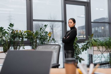 Young asian designer standing with crossed arms looking at laptop near blurred coffee to go in studio 