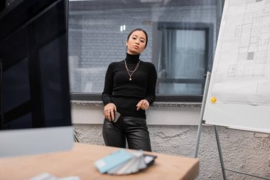 Asian interior designer holding smartphone near blurred computer and blueprint in office 