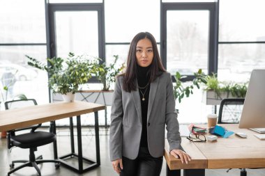 Brunette asian designer looking at camera near working table in studio  clipart