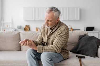 aged man with parkinson syndrome suffering from tremor in hands while sitting on sofa near walking cane