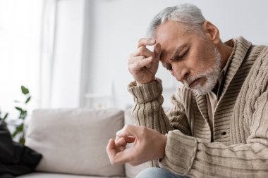 grey haired man with parkinson syndrome and tremor in hands sitting with closed eyes on couch at home