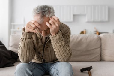 senior man with parkinson disease and trembling hands sitting with bowed head on couch at home