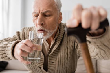 grey haired man with parkinson disease and trembling hands drinking water while sitting with blurred walking cane at home