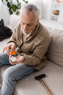 grey haired man suffering from parkinsonism and hands tremor holding pills container near walking cane on couch at home