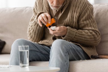 partial view of aged man with parkinson syndrome sitting near blurred glass of water and holding pills container in trembling hands