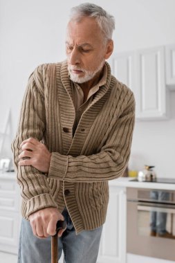 tensed man in knitted cardigan standing with walking cane in kitchen while suffering from parkinson disease clipart