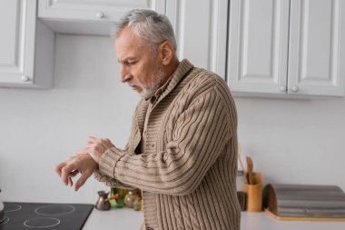 side view of man with with parkinson disease looking at trembling hands while standing in knitted cardigan in kitchen