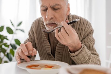 senior bearded man suffering from parkinsonism and holding spoon in trembling hand near plate with soup in kitchen